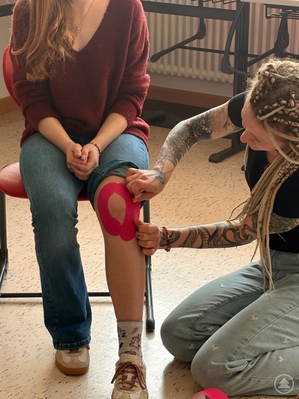 Eine Person zeigt einer Sch&uuml;lerin das Anlegen eines Kinesiotapes am Knie.