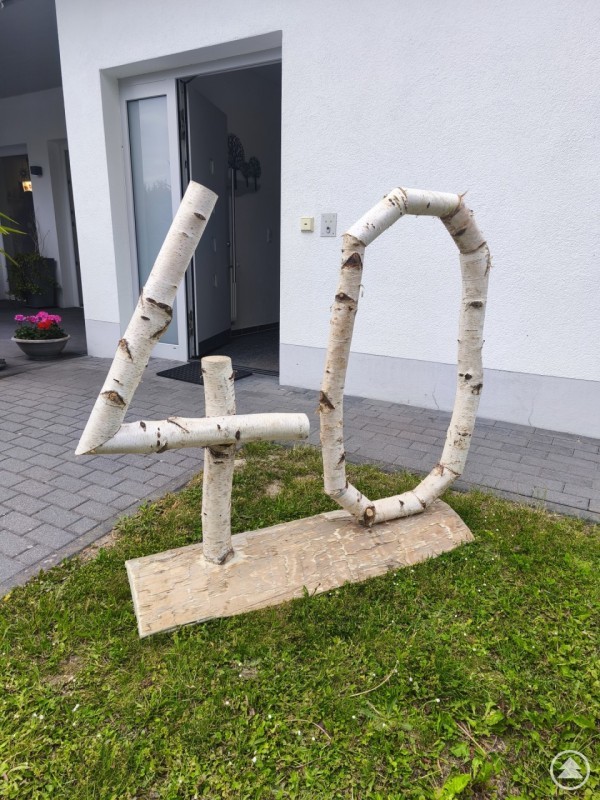 Das Bild zeigt eine große „40“, die aus weißen Birkenästen gefertigt wurde. Die Ziffern sind auf einem Holzbrett montiert und stehen auf einer kleinen Rasenfläche vor einem modernen Haus. Diese Art von Dekoration wird häufig für runde Geburtstage im Eingangsbereich verwendet – vermutlich handelt es sich hier also um eine Geburtstagsfeier zum 40. Geburtstag.