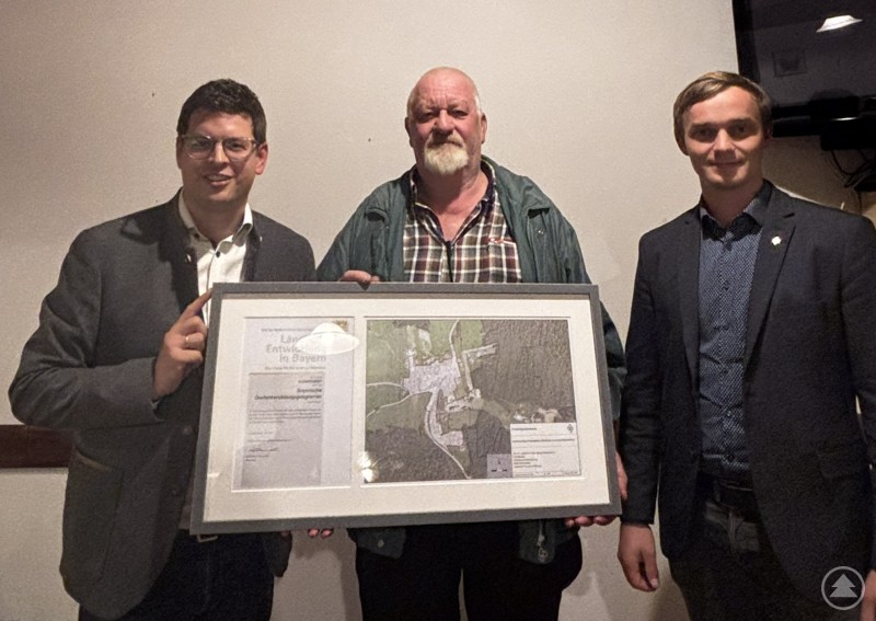 Bürgermeister Martin Pichler und Andreas Jobst freuten sich zusammen mit Michael Bartmaier (ALE Niederbayern), dass auch über Förderung an Privat ein gefälliges Dorfbild erreicht werden kann. Foto: Markt Schönberg Drei Männer stehen nebeneinander und halten gemeinsam einen großen gerahmten Plan mit einer Karte und einem offiziellen Dokument. Sie lächeln in die Kamera und befinden sich in einem Innenraum mit neutralem Hintergrund.