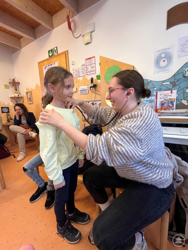 Eine Medizinstudentin untersucht ein Kindergartenkind mit einem Stethoskop, w&auml;hrend das Kind ruhig steht. Im Hintergrund sitzen weitere Personen in einem Gruppenraum.