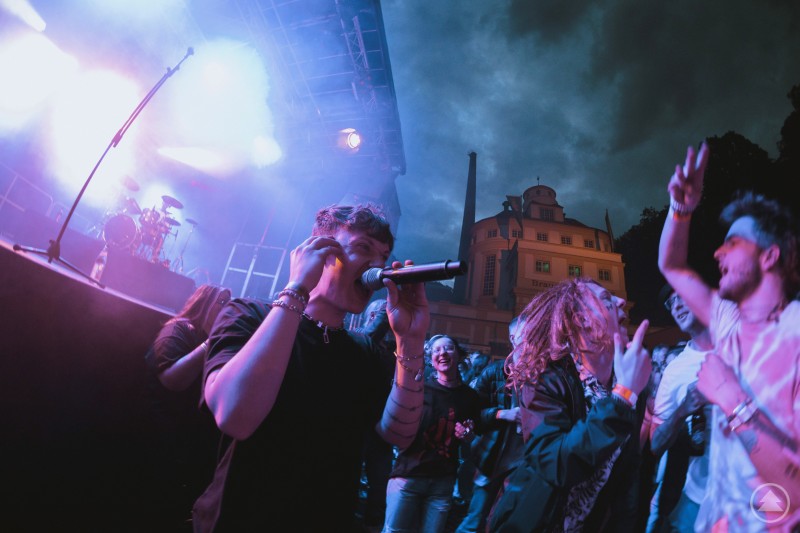 Ein junger Sänger steht mit Mikrofon vor einer feiernden Menschenmenge bei einem Open-Air-Konzert. Die Bühne im Hintergrund ist in lila-blaues Licht getaucht, während das Publikum ausgelassen mitfeiert. Im Hintergrund ist ein historisches Gebäude mit der Aufschrift 