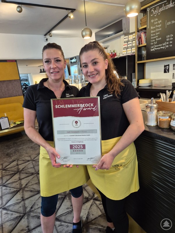 Zwei Frauen in schwarzem Oberteil und gelber Schürze stehen in einem modernen Café. Sie halten gemeinsam eine Urkunde mit der Aufschrift „Schlemmerblock Award 2025“ in die Kamera und lächeln.