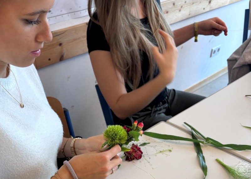 Beim Floristen entstanden liebevoll und kreativ gestaltete kleine Gestecke. Zwei junge Frauen sitzen an einem Tisch und gestalten florale Gestecke mit Blumen und Gräsern.