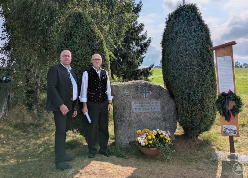 Bürgermeister Roland Schraml und Theodor Bauer am Gedenkstein in Haidmühle. Zwei Männer in traditioneller Tracht stehen neben einem Gedenkstein, der mit Blumen geschmückt ist. Rechts daneben befindet sich ein Holzständer mit Tafel und Kranz, im Hintergrund sind Bäume und eine Wiese zu sehen.