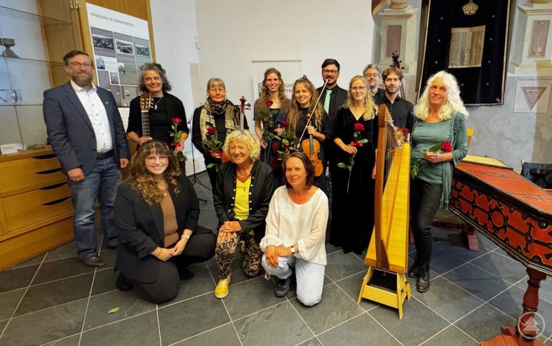Die Organisatoren der Freundschaftskonzerte (links) Sachgebietsleiter Christian Fiebig, Kulturreferentin Gabriela Rauscher (vorne links) vom Landkreis Freyung-Grafenau sowie Hana Dědová (vorne rechts) von der Synagoge Čkyně freuen sich zusammen mit der stellvertretenden Landrätin Hilde Greiner (vorne mittig) und dem Ensemble EnsaladaMixta in Čkyně über einen gelungenen Konzertabend. Eine Gruppe von Musikerinnen und Musikern sowie Organisatoren posiert nach einem Konzert in einem historischen Saal. Einige halten Rosen in der Hand, im Hintergrund sind Instrumente wie Harfe und Gitarre zu sehen.
