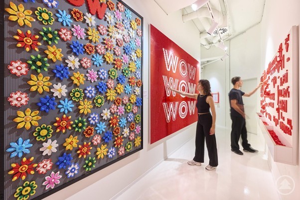 In einem hellen Galerieraum h&auml;ngen gro&szlig;formatige, bunte Kunstwerke: links ein Mosaik aus vielen farbenfrohen Blumen, in der Mitte ein rotes Bild mit der Aufschrift &bdquo;WOW WOW WOW&ldquo;. Zwei Personen betrachten die Ausstellung, rechts ist eine Wand mit roten Schrift-Elementen zu sehen.