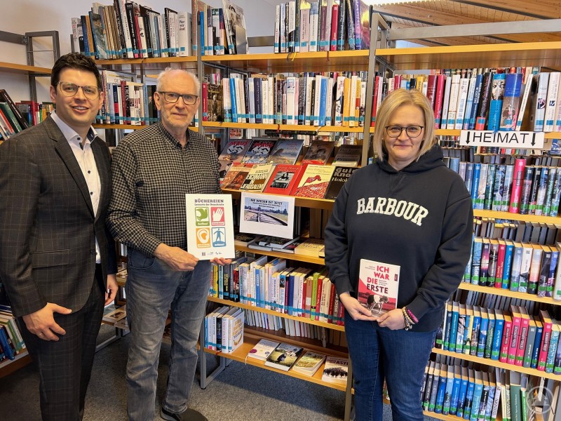Bürgermeister Martin Pichler (v.l.), Reinhold Hartl und Gemeinderätin Carolin Garhammer freuen sich bewusst in einer Ecke zum Thema Geschichte und Diktatur über das Siegel - und noch mehr darüber, dass Büchereien als wirksames Mittel gegen Fake-News weiterhin wirken. Drei Personen stehen in der Marktbücherei Schönberg vor Bücherregalen. Eine Person hält ein Zertifikat in der Hand, eine andere ein Buch.