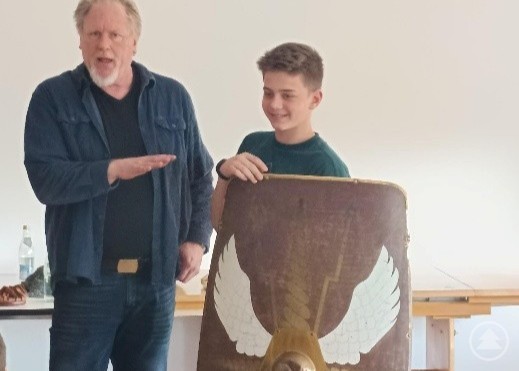 Bei einem spannenden Vortrag von Bernhard Bischler über die Römerzeit lernten die Realschüler, wie wichtig die richtige Ausrüstung für die langen Märsche der Legionen war. Ein Archäologe erklärt einem Schüler eine römische Ausrüstung, während dieser ein großes Schild hält.