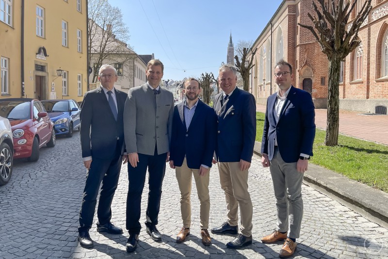Kamen zur Mitgliederversammlung nach Landshut: Prof. Dr. Günter Dippold, stellvertretender Vorsitzender des Landesvereins für Heimatpflege und Bezirksheimatpfleger Oberfranken (von links), Bezirkstagspräsident und Vorsitzender des Landesvereins Dr. Olaf Heinrich, Schatzmeister des Landesvereins Jun.-Prof. Dr. Johannes Lorenz, Geschäftsführer des Landesvereins Dr. Rudolf Neumaier und der niederbayerische Bezirksheimatpfleger Dr. Clemens Knobling. Das Bild zeigt fünf Männer in Anzug oder Tracht, die bei sonnigem Wetter auf einer gepflasterten Straße vor historischen Gebäuden posieren. Im Hintergrund ist eine Kirche mit Backsteinfassade sowie der Turm einer weiteren Kirche zu erkennen – vermutlich ein bekannter Ort in Bayern. Die Szenerie wirkt offiziell, etwa im Rahmen eines Treffens, Pressetermins oder politischen Austauschs. Alle Personen blicken lächelnd in die Kamera, was auf eine positive Stimmung und ein erfolgreiches Treffen schließen lässt.