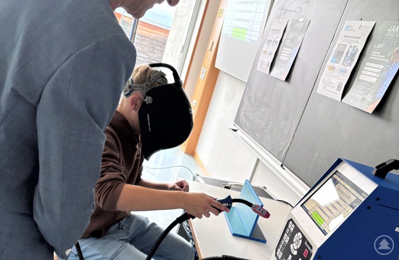 Eine Person trägt eine Schweißschutzbrille und arbeitet an einem modernen Schweißsimulator, ein älterer Mann beobachtet.