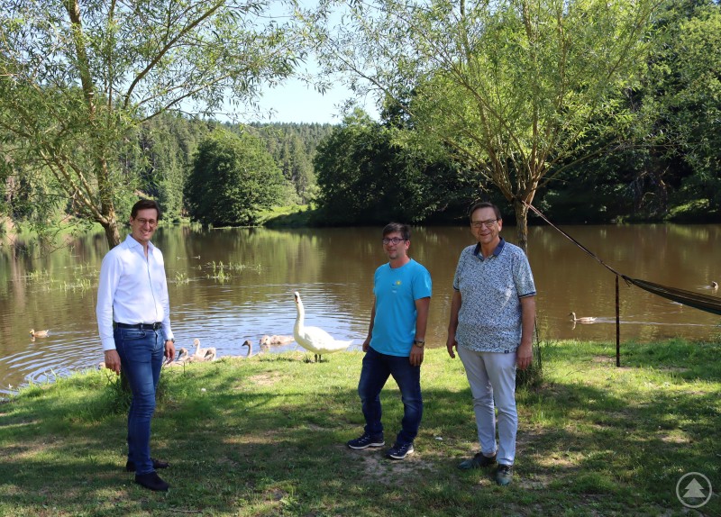 MdL Stefan Ebner (l.) und Bürgermeister Franz Wittmann (r.) zeigten sich begeistert von der Campinganlage Höllensteinsee in Pirka, die ihnen vom Betreiber Hans Müller vorgestellt wurde. Drei Männer stehen am Ufer des Höllensteinsees, umgeben von Bäumen und Wasser. Im Hintergrund schwimmen Schwäne und Enten.