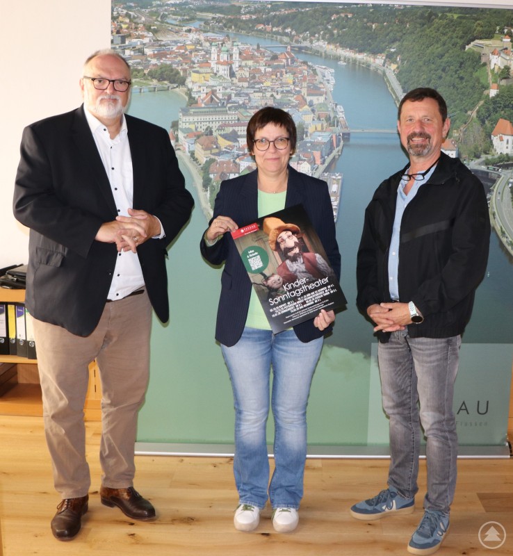 Vorstellung des Programms für das Kindersonntagstheater 2025/2026 im Jugendzentrum Zeughaus. Drei Personen stehen vor einem großen Wandbild der Stadt Passau. Die Frau in der Mitte hält ein Plakat für das Kindersonntagstheater 2025/2026 in den Händen. Links und rechts von ihr stehen zwei Männer, alle drei lächeln in die Kamera.