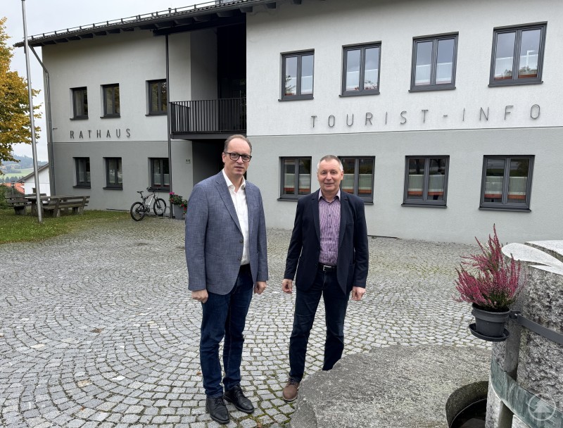 MdL Martin Behringer (l.) besuchte Bürgermeister Robert Muhr (r.) im Rathaus Kirchberg, um über aktuelle kommunale Themen und Herausforderungen zu sprechen. Zwei Männer stehen vor dem modernen Rathaus- und Tourist-Info-Gebäude in Kirchberg im Wald. Beide tragen Sakkos, im Hintergrund sind Fahrräder, eine Bank und herbstlich gefärbte Bäume zu sehen.