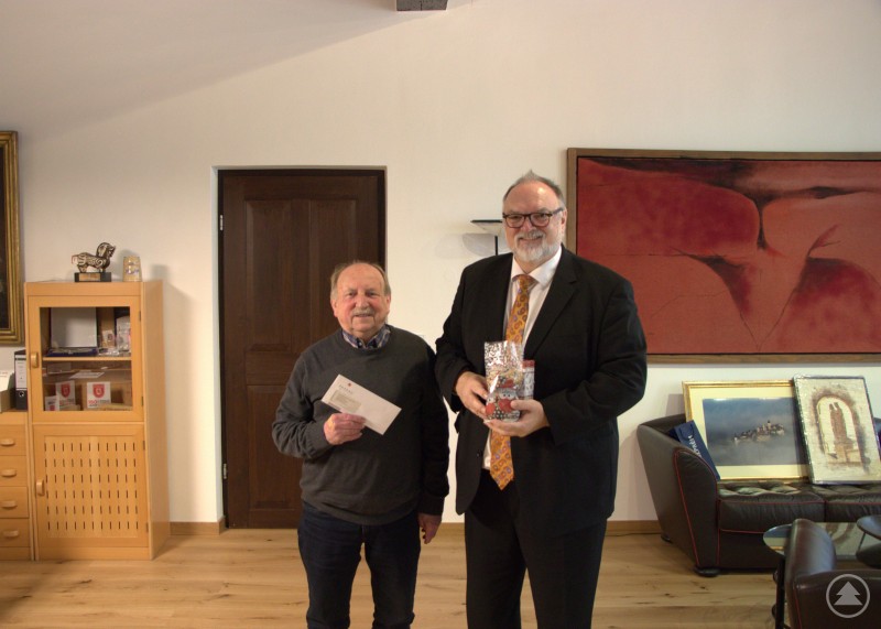 Oberbürgermeister Jürgen Dupper (rechts) mit dem Vorstand der Passauer Dreiflüssemusikanten Hermann Kapfhammer. Zwei Männer stehen in einem Büro und lächeln in die Kamera. Einer hält ein Kuvert, der andere ein verpacktes Geschenk.