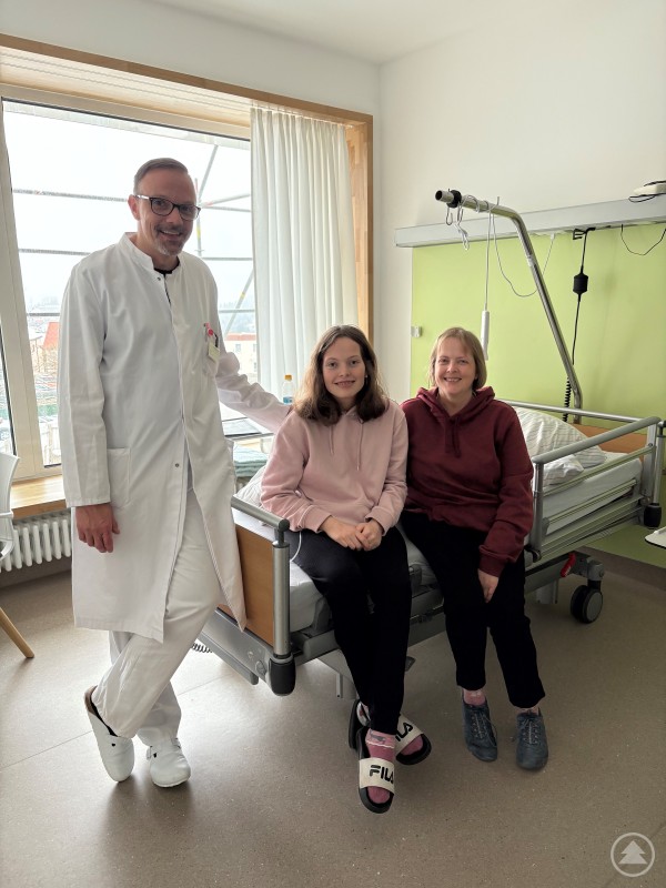 v.l.: Chefarzt Kay Seidel, Patientin Frau Freitag und deren Mutter freuen sich über die Ergebnisse der Operation. Ein Arzt im weißen Kittel steht neben einer jungen Patientin und deren Mutter, die gemeinsam auf einem Krankenhausbett sitzen. Alle drei lächeln in einem freundlich wirkenden Patientenzimmer in die Kamera.