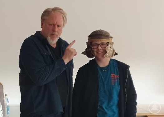 Ein Sch&uuml;ler tr&auml;gt einen r&ouml;mischen Helm und l&auml;chelt, w&auml;hrend der Referent daneben steht und etwas erkl&auml;rt.