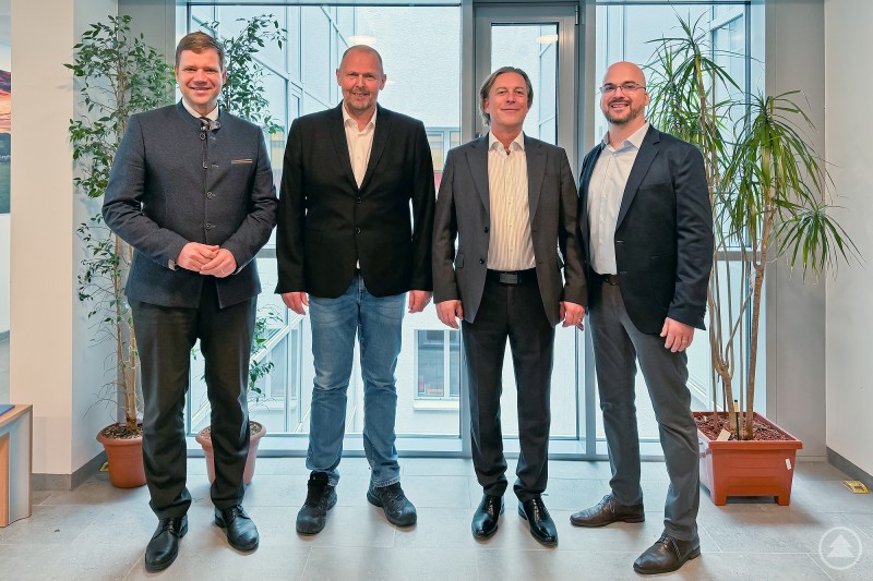 Bezirkstagspräsident Dr. Olaf Heinrich (von links), Walter Blüml, Prof. Dr. Joachim Nitschke und Dr. Philipp Holter. Vier Männer in Businesskleidung stehen nebeneinander in einem modernen Gebäude mit großen Fenstern und Pflanzen im Hintergrund und blicken in die Kamera.