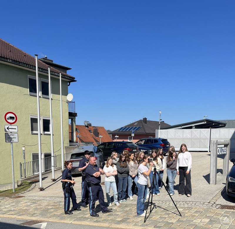 Unter Aufsicht des Verkehrsspezialisten der PI Freyung POK Christian Moosbauer sowie POW Marina Blöchl und POW Alexander Weiß konnten die Schülerinnen einen Blick durch die Kamera zur Geschwindigkeitsmessung werfen. Das Bild zeigt eine Gruppe Jugendlicher vor einer Polizeistation bei sonnigem Wetter. Zwei Polizisten stehen mit der Gruppe, während ein Mädchen hinter einer Kamera auf einem Stativ steht – vermutlich wird gerade ein Video gedreht oder ein Foto gemacht. Die Szene wirkt wie ein Projekttag, ein Besuch im Rahmen der schulischen Berufsorientierung oder ein medienpädagogisches Projekt mit Beteiligung der Polizei. Im Hintergrund sind geparkte Autos, ein Gebäudekomplex mit „POLIZEI“-Schild sowie Wohnhäuser mit Solaranlagen zu sehen. Die Stimmung scheint konzentriert und freundlich.