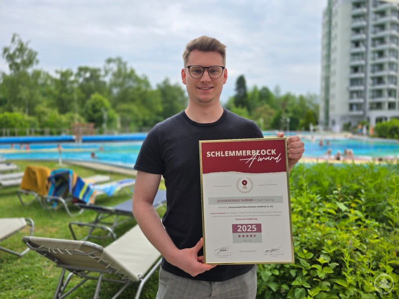 Florian Sengstock, Marketing Manager der Johannesbad Therme Bad Füssing, freut sich über den Schlemmerblock-Award 2025 in der Kategorie Freizeit für die Region Passau & Umgebung. Ein junger Mann steht vor einem Außenbecken der Therme und hält eine gerahmte Urkunde mit dem Titel „Schlemmerblock Award 2025“. Im Hintergrund Liegestühle, Badegäste und ein mehrstöckiges Gebäude.