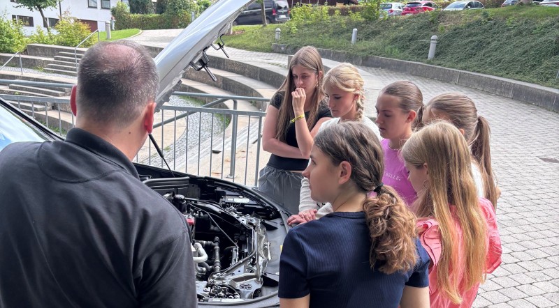 Eine Gruppe Jugendlicher steht um einen geöffneten Motorraum eines Autos, ein Mann erklärt technische Details.