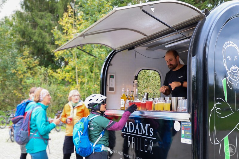 Überraschung auf der Strecke: Die „Cocktailerie Ädäm’s“ versorgte die Radler mit fruchtigen Drinks. Ein Mann bereitet bunte Getränke in einem mobilen Cocktailtrailer zu, während Radfahrerinnen in Sportkleidung ihre Getränke entgegennehmen.