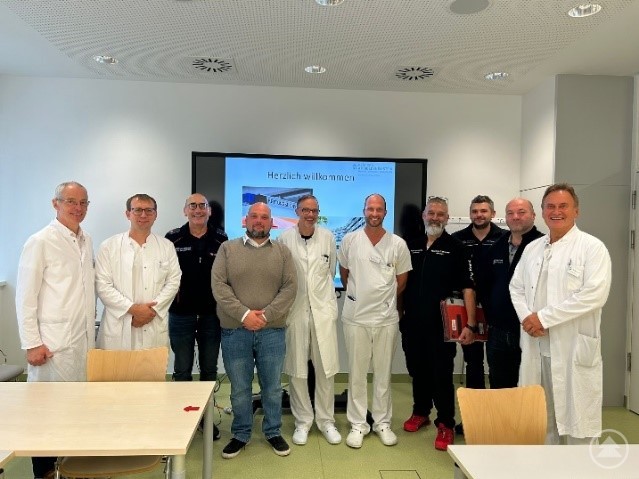 Vertreter der Rettungsdienste sowie Ärzte und Mitarbeiter der Kliniken Am Goldenen Steig beim Austausch im Krankenhaus Freyung. Ärzte und Vertreter der Rettungsdienste stehen gemeinsam in einem Konferenzraum des Krankenhauses Freyung, im Hintergrund läuft eine Präsentation über die Notfallversorgung.