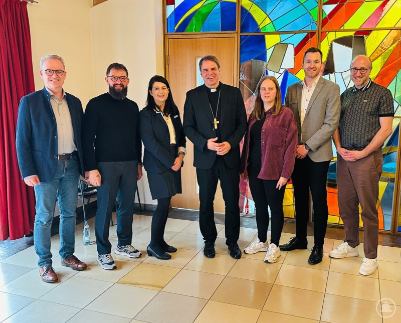 Stefan Seiderer (Bischöflich Beauftragter), Wolfgang Gaßler (Fachgebietsleitung), Alexandra Aulinger-Lorenz (hauptberufliche Vorständin), Bischof Stefan Oster SDB, Maria Petzi (stv. Fachgebietsleitung), Florian Kasparak (geschäftsführender Vorstand) und Dr. Peter Robl (Aufsichtsratsvorsitzender) Sieben Personen stehen vor einem farbigen Kirchenfenster für ein gemeinsames Gruppenfoto.