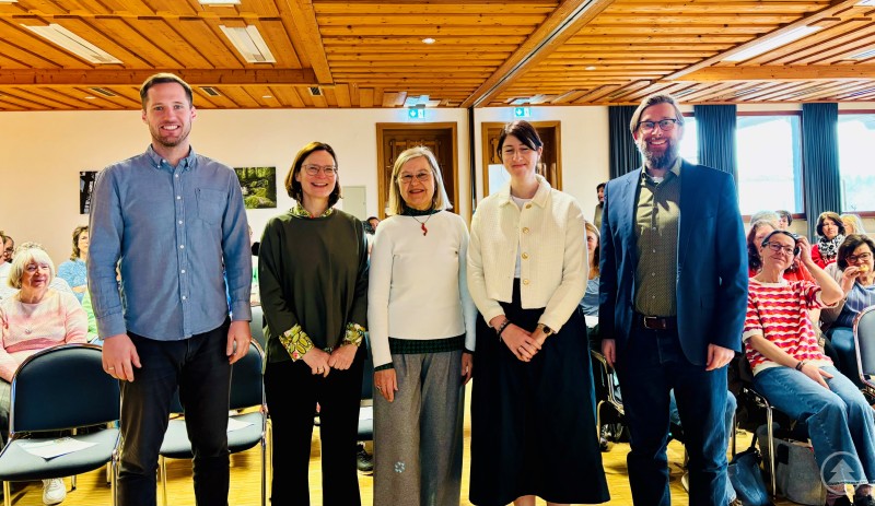 Freuten sich über das große Interesse an der Schulung (von links) René Kurtz (Gesundheitsregion Plus), Susanna Kurka, Johanna Myllymäki (beide von der Fachstelle Pflege), Nicole Polleichtner (Kreisseniorenbeauftragte) und Christian Fiebig (Sachgebietsleiter Landkreisangelegenheiten). Das Bild zeigt eine Gruppe von fünf Personen, die in einem hellen Veranstaltungssaal vor einem größeren Publikum stehen und freundlich in die Kamera lächeln. Die Szenerie wirkt wie eine offizielle Schulung oder Fortbildung – im Hintergrund sitzen zahlreiche Teilnehmende, überwiegend Frauen, die dem Geschehen aufmerksam folgen. Die Holzdecke und Einrichtung deuten auf eine ländlich geprägte Location hin, möglicherweise ein Bürgerhaus oder Gemeindesaal. Die Atmosphäre ist offen, engagiert und von gegenseitigem Interesse geprägt – passend zu einer Veranstaltung im sozialen oder ehrenamtlichen Bereich.