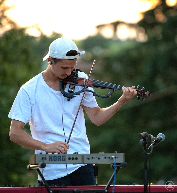 Ein junger Mann spielt eine elektrische Geige und bedient gleichzeitig ein KORG-Keyboard, im Freien bei Sonnenuntergang.