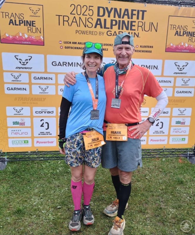 Ein Vater-Tochter-Team steht nach dem Zieleinlauf des Transalpine Run vor der offiziellen Sponsorenwand, beide tragen ihre Finisher-Medaillen und Startnummern und strahlen in die Kamera.