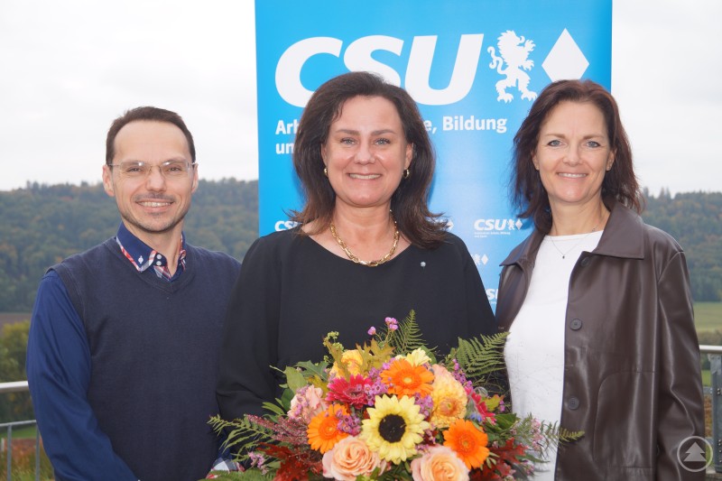 Dr. Roland J. Kufner, Dr. Ute Eiling-Hütig und Waltraud Eder bei der Landesversammlung des AKS der CSU Bayern in Beilngries. Drei Personen stehen vor einem CSU-Banner im Freien. Die mittlere Person hält einen bunten Blumenstrauß mit Dahlien und Herbstblumen. Alle drei lächeln in die Kamera, im Hintergrund ist bewaldete Landschaft zu sehen.