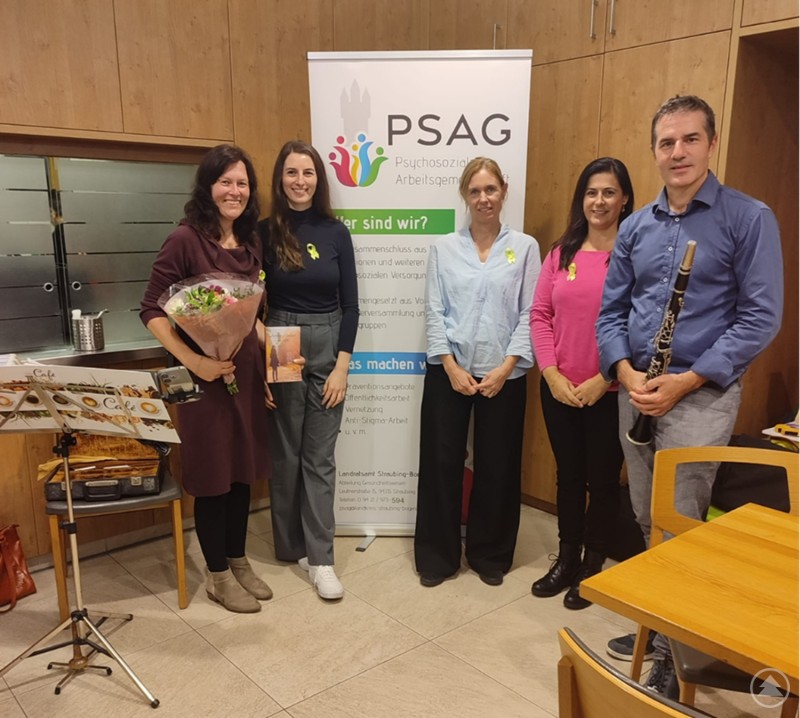 Nicole Lindner (Referentin, 2. v. l.) mit ihrem Ehemann (r.) sowie Christina Drexler (PSAG, Mitte), Melanie Dullinger (Familienhaus Christuskirche) und Luisa Hammerschmied (PSAG, 2. v. r.) bei der Lesung zum Tag der seelischen Gesundheit. Fünf Personen stehen lächelnd vor einem Banner mit der Aufschrift „PSAG – Psychosoziale Arbeitsgemeinschaft“. Eine Frau hält einen Blumenstrauß, ein Mann eine Klarinette. Die Gruppe befindet sich in einem Raum mit Holzmöbeln und freundlicher Atmosphäre.