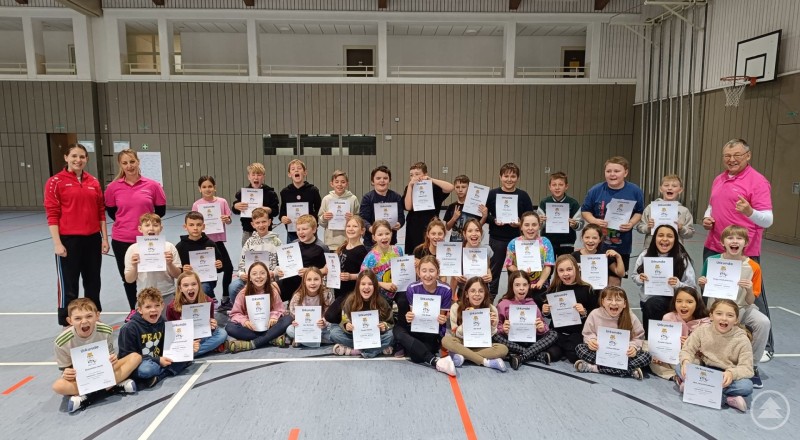 Viertklässler der GS Spiegelau präsentieren stolz ihre Urkunden. Gruppenfoto in einer Turnhalle. Viertklässler der Grundschule Spiegelau sitzen und stehen in mehreren Reihen und halten Urkunden in die Kamera. Links und rechts stehen Betreuerinnen und ein Trainer.