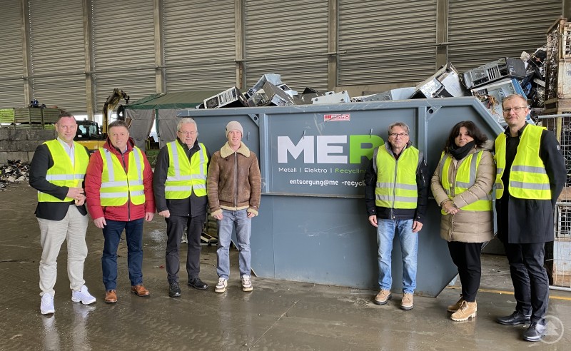 Michael Prietsch und Daniela Königbauer (rechts) beim Besuch der Firma MER in Oberschneiding. Sieben Personen in Warnwesten stehen in einer Halle neben einem Container voller ausgedienter Elektrogeräte, im Hintergrund Altmetall und Maschinen.