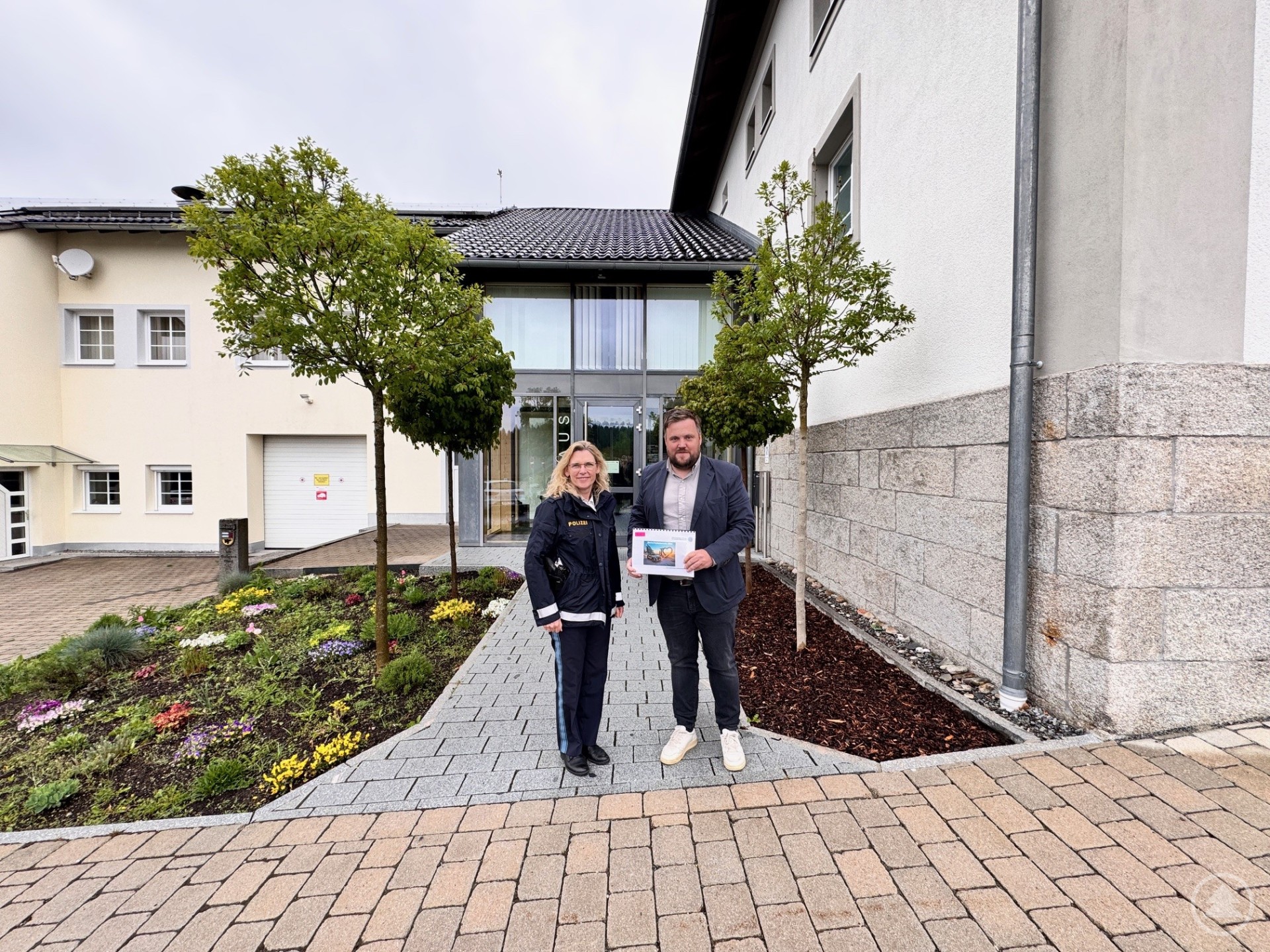 Eine Frau und ein Mann stehen vor dem Eingang eines modernen Gebäudes, der Mann hält ein Dokument in der Hand. Neben ihnen zwei kleine Bäume und ein gepflegtes Blumenbeet.