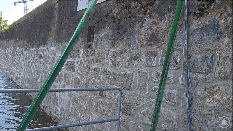 Sanierte Donaukaimauer Die sanierte Ufermauer an der Donau in Passau zeigt frisch verfugtes Natursteinmauerwerk. Eine Absperrung mit grünen Gurten und ein Geländer sichern die Baustelle. Die Mauer verläuft entlang des Wassers, im Hintergrund sind Baukräne und ein Schild zu sehen.