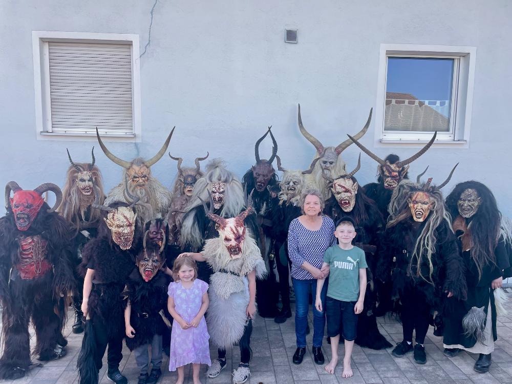 Eine Gruppe von Perchten und Krampussen mit gruseligen Masken und Hörnern posiert gemeinsam mit mehreren Kindern und Erwachsenen vor einer Hauswand.