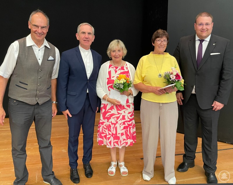 Fünf Personen stehen auf einer Bühne, zwei Frauen in der Mitte halten Blumensträuße in den Händen, daneben stehen drei Männer in festlicher Kleidung.