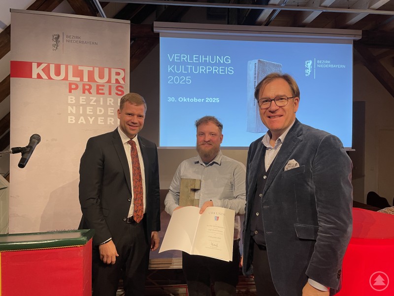 Dr. Olaf Heinrich (l.) und Dr. Thomas Pröckl (r.) überreichten den Kulturpreis des Bezirks Niederbayern an Christian Schwarz, den Leiter des Jugendcafé Zwiesel. Drei Männer stehen bei einer Preisverleihung auf einer Bühne, einer von ihnen hält eine Urkunde und eine Skulptur. Im Hintergrund ist eine Leinwand mit der Aufschrift „Verleihung Kulturpreis 2025“ zu sehen.