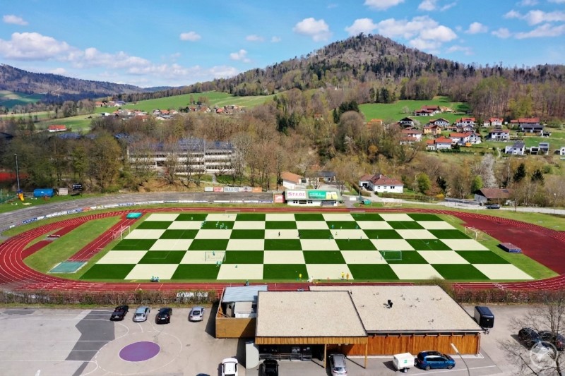Das Stadion in Hauzenberg wurde symbolisch in ein Schachbrett verwandelt. Luftaufnahme eines Sportgeländes mit Laufbahn und Fußballfeld, das in einem auffälligen Schachbrettmuster angelegt ist.