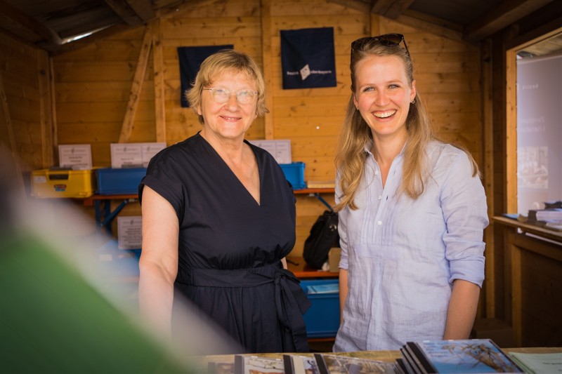 Zwei Frauen stehen l&auml;chelnd in einem Holzstand und schauen in die Kamera. Vor ihnen liegen B&uuml;cher oder Brosch&uuml;ren aus, im Hintergrund sind Regale und Infomaterial zu erkennen.
