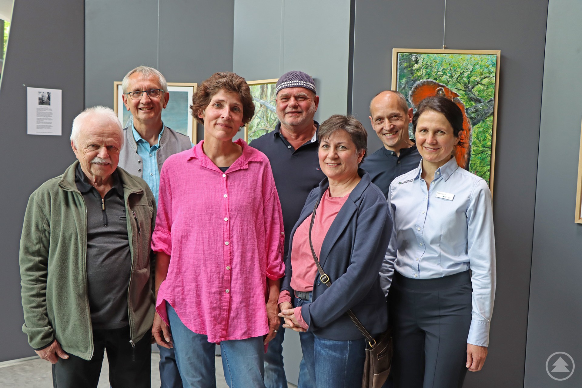 Sieben Personen stehen lächelnd vor einer Wand mit farbenfrohen Kunstwerken in einer Ausstellung.
