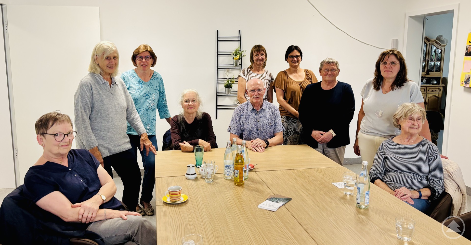 Elf Personen, davon einige sitzend, andere stehend, versammeln sich um einen Besprechungstisch in einem hellen Raum mit Getränken und Gläsern auf dem Tisch.