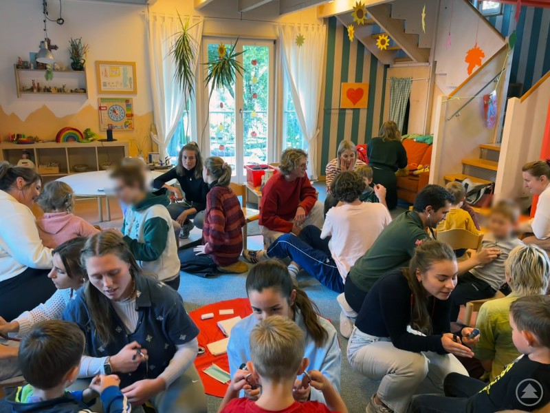 Die angehenden Medizinerinnen und Mediziner beim Untersuchen der Kinder. Mehrere Studierende sitzen gemeinsam mit Kindern auf dem Boden eines bunt dekorierten Gruppenraums und führen spielerisch Untersuchungen durch. Die Atmosphäre ist lebendig und freundlich.