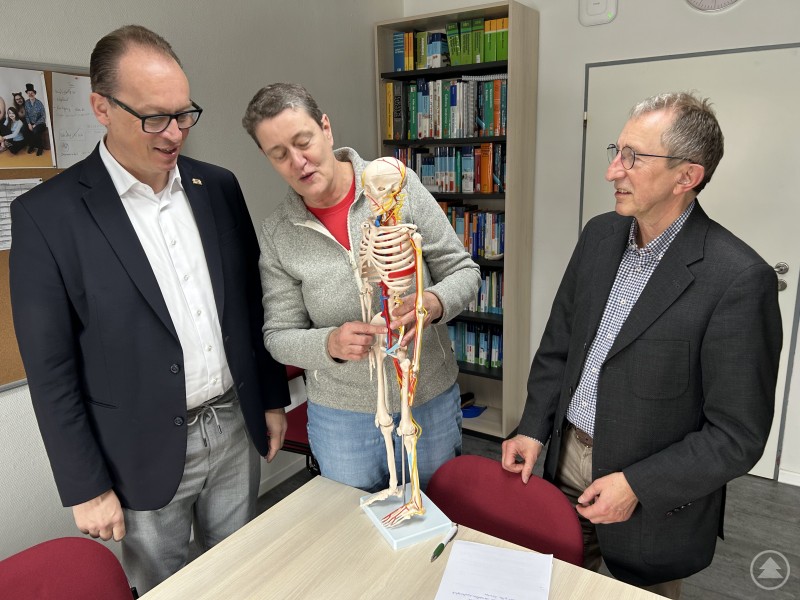 Wie bei einem Körper spielen auch für eine Schule viele Faktoren zum Gelingen zusammen. MdL Martin Behringer (v.l.) zeigte sich sehr interessiert daran. Schulleiterin Astrid Schricker und Geschäftsführer Ludwig Brunner bedankten sich für den Besuch des Abgeordneten. Drei Personen stehen gemeinsam an einem Tisch in einem Klassenraum oder Büro. In der Mitte wird ein anatomisches Skelettmodell betrachtet und erklärt. Im Hintergrund sind Bücherregale mit medizinischer Fachliteratur zu sehen. Alle drei Personen wirken interessiert und im Gespräch vertieft.