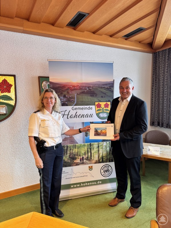 PORin Ingrid Grötzinger übergibt Bürgermeister Josef Gais den Sicherheitsbericht der Gemeinde Hohenau. Eine Polizeibeamtin in Uniform und ein Bürgermeister in Anzug stehen in einem Besprechungsraum vor einem Roll-up der Gemeinde Hohenau. Die Beamtin überreicht ein Dokument mit einem Titelblatt an den Bürgermeister. Beide lächeln in die Kamera.