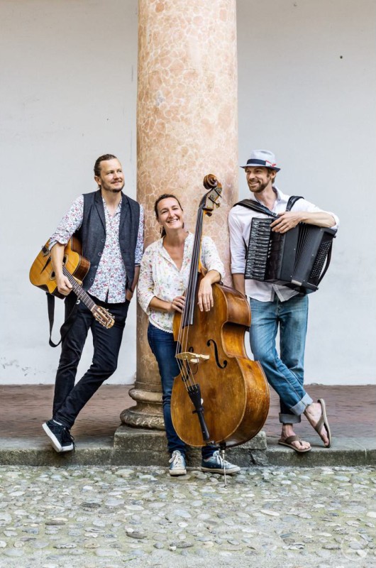 Drei Musiker posieren entspannt mit Kontrabass, Gitarre und Akkordeon vor einer Säule im Freien.