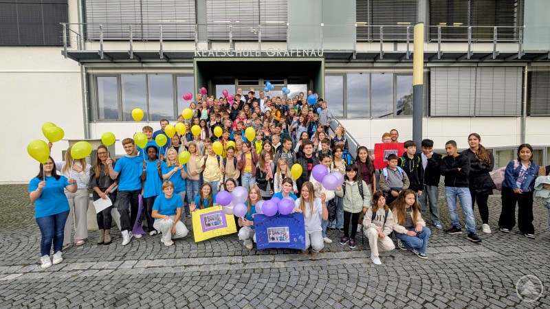 Konrektor Martin Weiß sowie die Tutorinnen und Tutoren begrüßten die neuen Fünftklässler und ihre Eltern an der Realschule Grafenau. Vor dem Eingang der Realschule Grafenau stehen zahlreiche neue Fünftklässler mit bunten Luftballons, begleitet von Tutoren in blauen T-Shirts. Alle wirken fröhlich und gespannt.