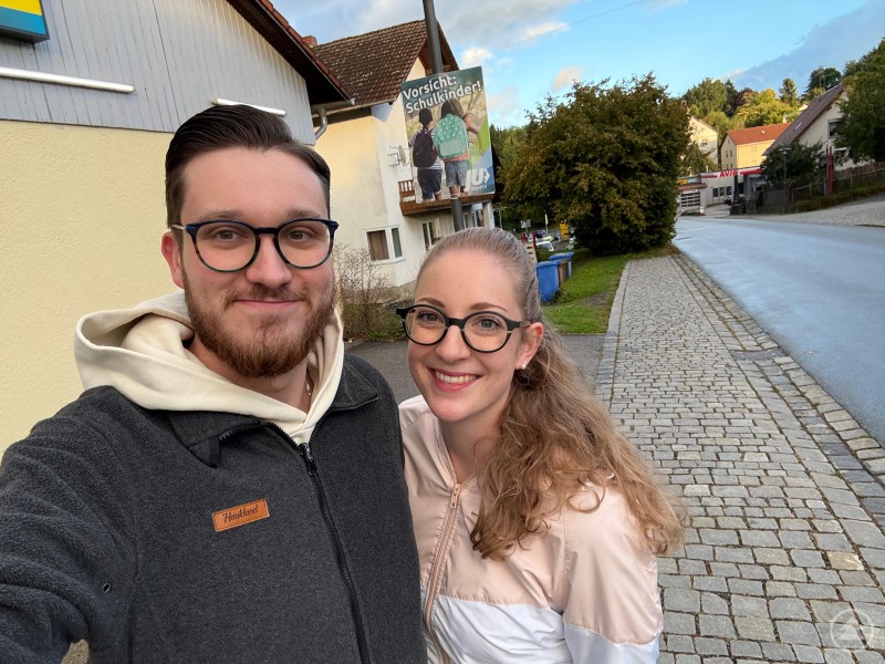 Ein junges Paar steht lächelnd vor einem Haus mit aufgehängtem Plakat „Vorsicht: Schulkinder!“.