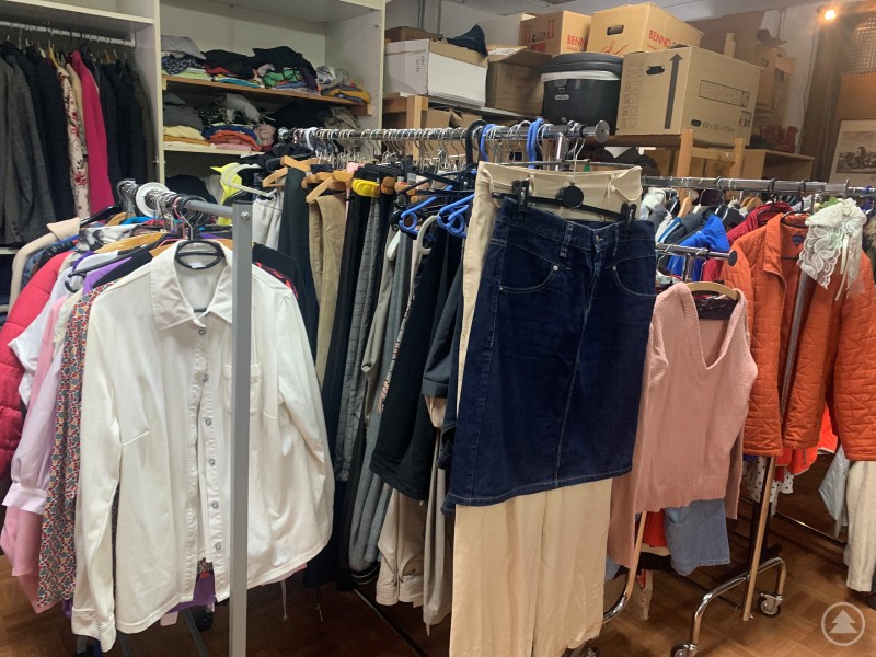 Klamotten in allen Größen, für Kinder und Erwachsene sowie Schuhe und vieles mehr findet sich in der Kleiderkammer des Aidenbacher Helferkreises. Kleiderständer mit Hemden, Hosen, Jacken und Pullovern in verschiedenen Farben und Größen stehen dicht aneinandergereiht in einem Raum. Im Hintergrund sind Regale mit gefalteter Kleidung und Kartons zu sehen.